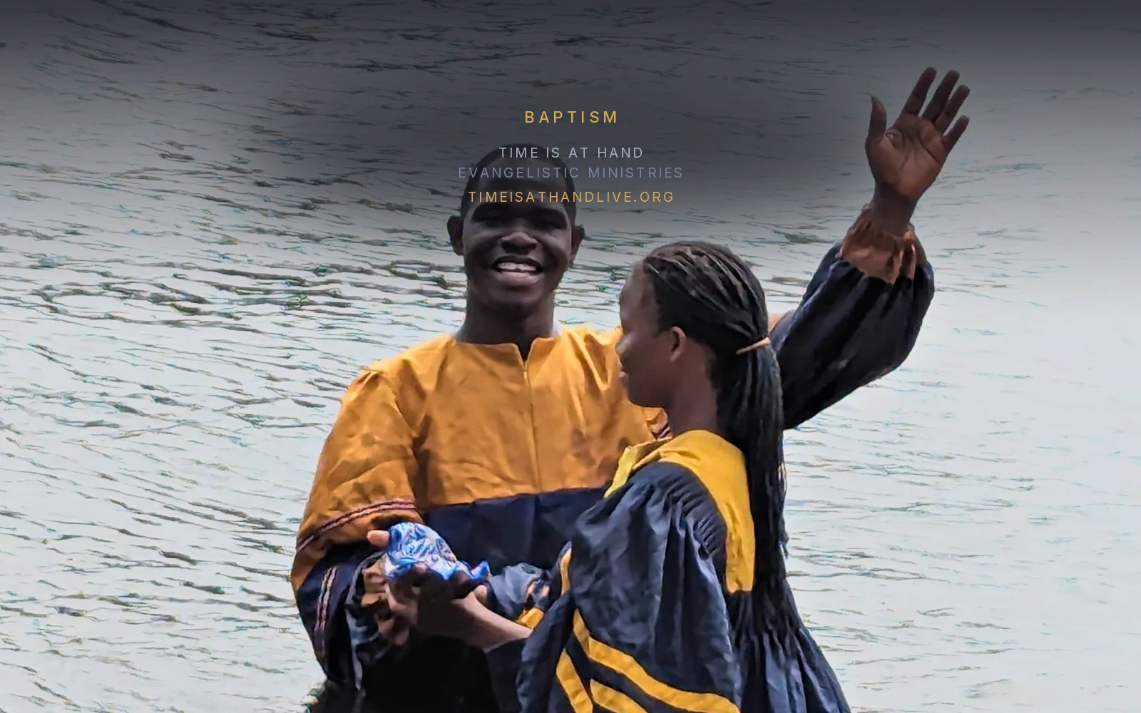 A minister in the river holding a candidate in a navy and gold baptismal gown, his hand raised as he speaks