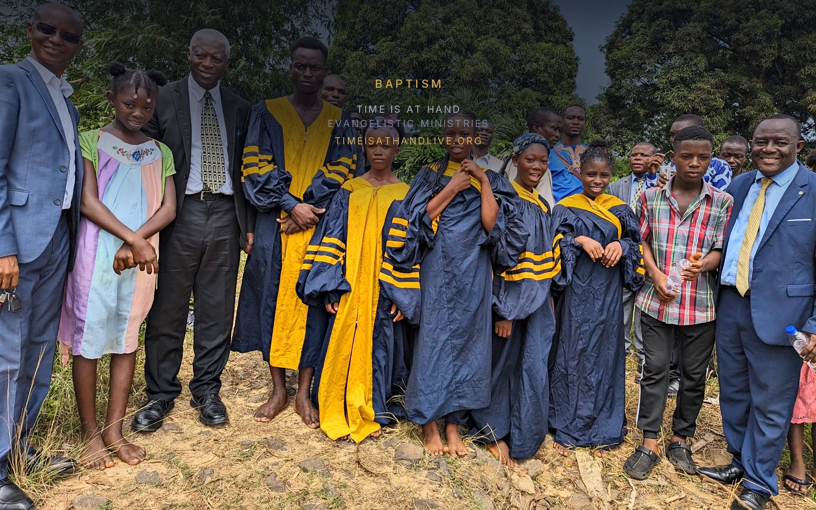 The baptismal candidates in navy and gold gowns standing barefoot on the stones with the ministers, trees behind them