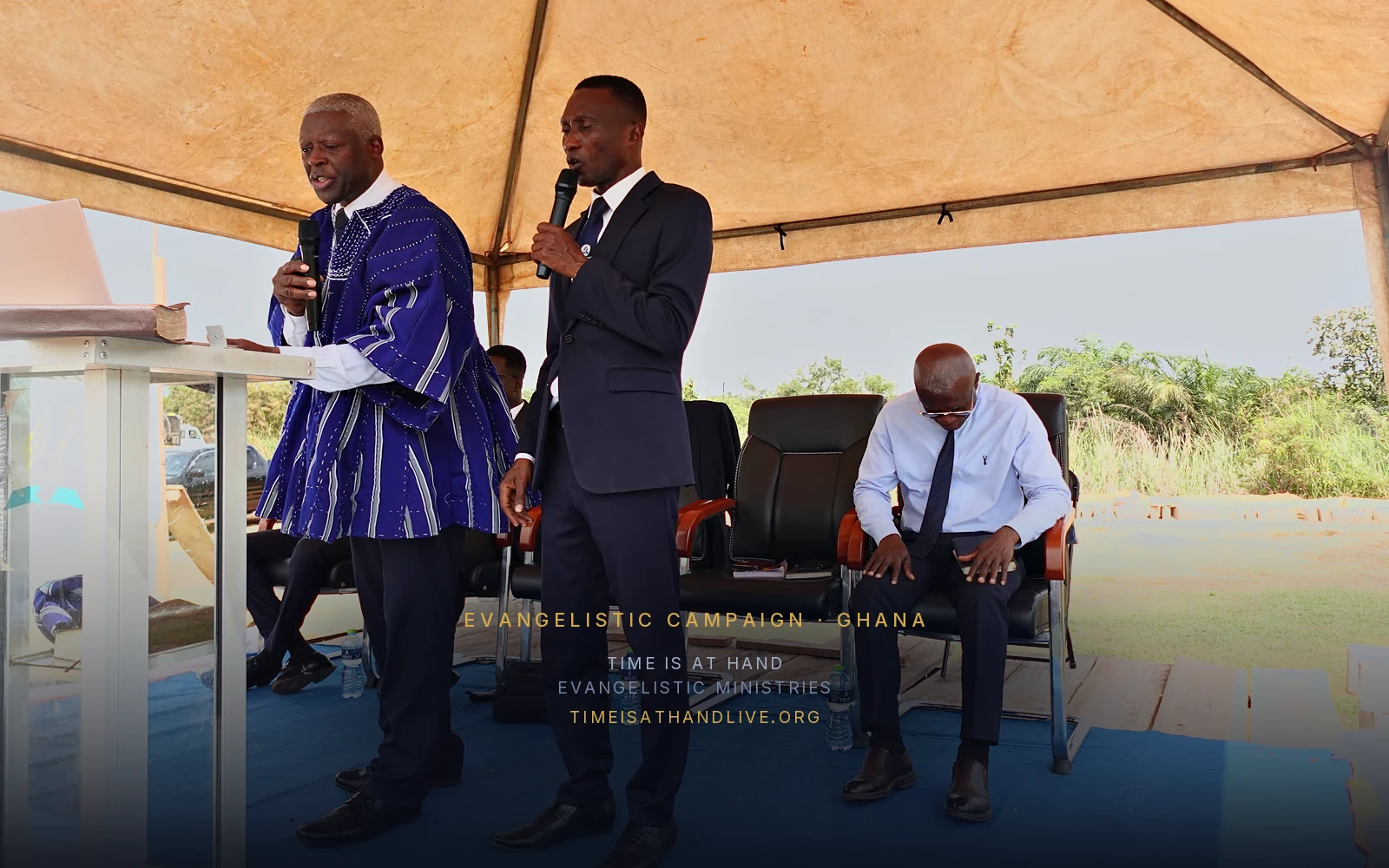 Two speakers at the pulpit under the canopy, a third seated beside them
