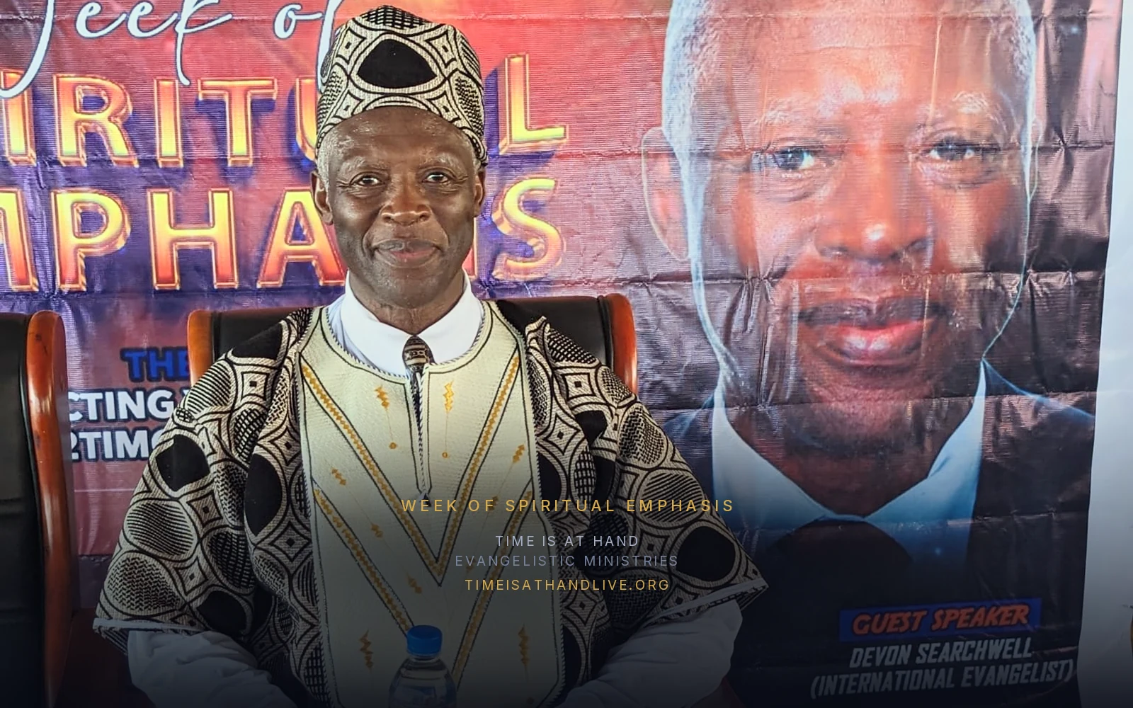 Devon Searchwell in a patterned West African robe and cap, seated beneath the Week of Spiritual Emphasis banner that names him guest speaker